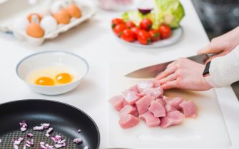 Mesa branca com alimentos, do fundo para a frente: uma caixa de ovos brancos e marrons aberta, um prato com tomate e alface, uma vasilha com dois ovos crus, uma panela preta com temperos dentro e uma mão branca feminina cortando carne em cubos sobre uma tábua branca de carne.