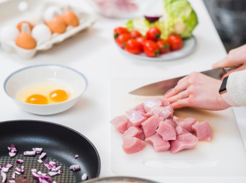 Mesa branca com alimentos, do fundo para a frente: uma caixa de ovos brancos e marrons aberta, um prato com tomate e alface, uma vasilha com dois ovos crus, uma panela preta com temperos dentro e uma mão branca feminina cortando carne em cubos sobre uma tábua branca de carne.