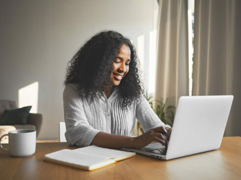 Mulher negra, jovem, de cabelos encaracolados, digita o teclado de um notebook, sorrindo e sentada em mesa com uma caneca e um caderno.