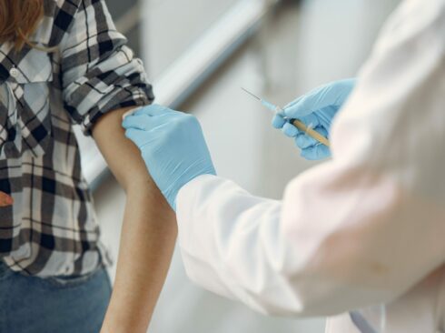 Mulher com camisa xadrez e jeans recebe vacina no braço de um profissional da saúde vestindo jaleco branco e luvas azuis