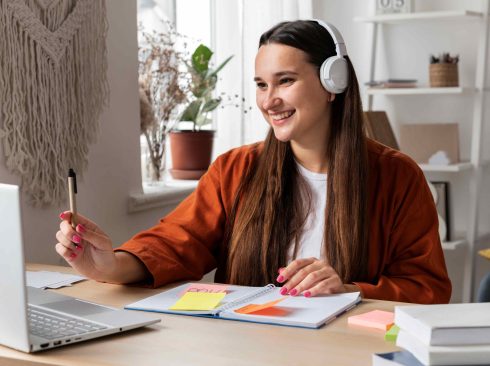 Mulher sorrindo em frente a um notebook, e na mesa alguns cadernos de anotação.